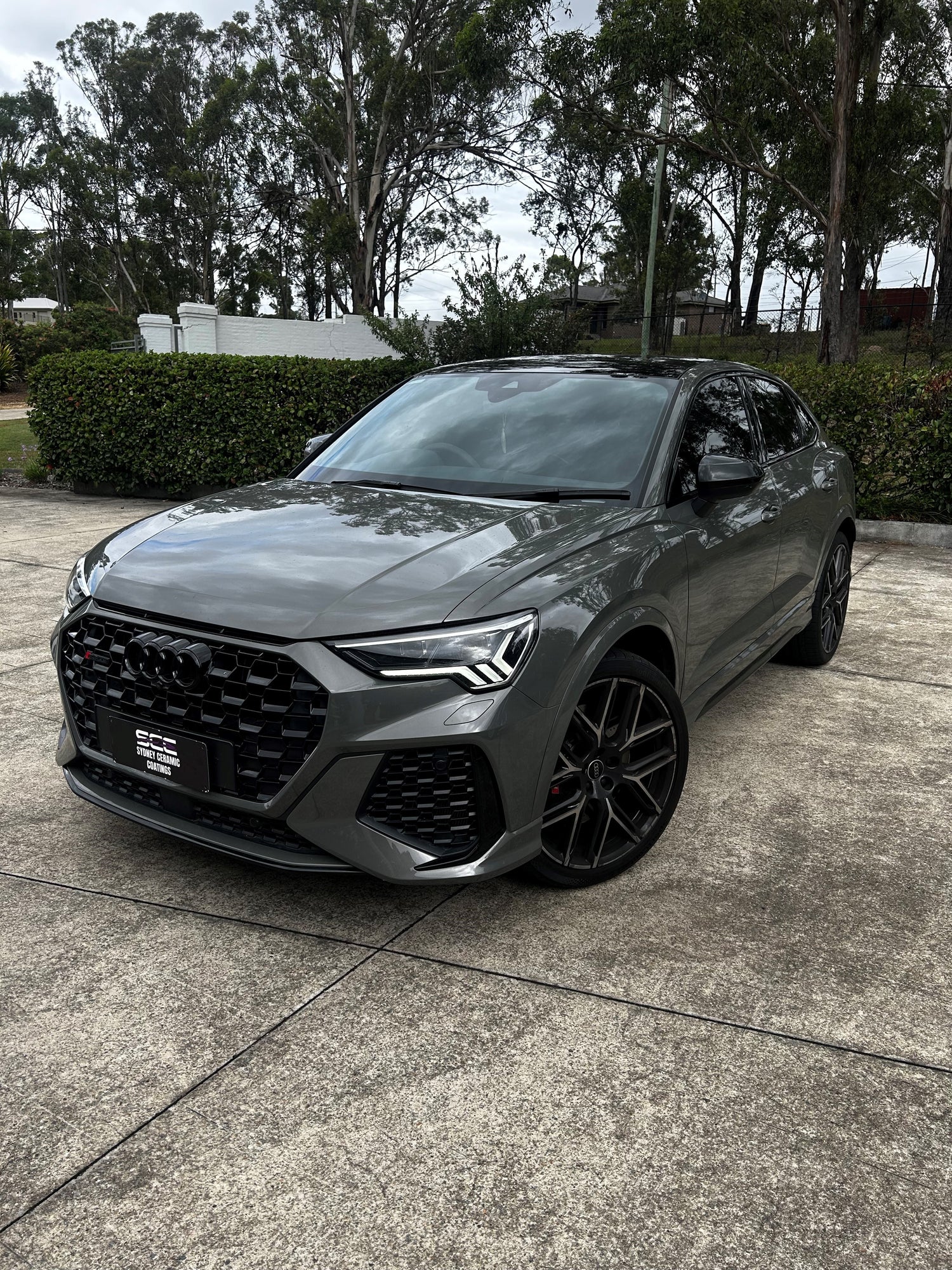 Grey Audi RS Q3 SUV with black wheels and aggressive styling, parked on a concrete surface near trimmed hedges and tall trees.