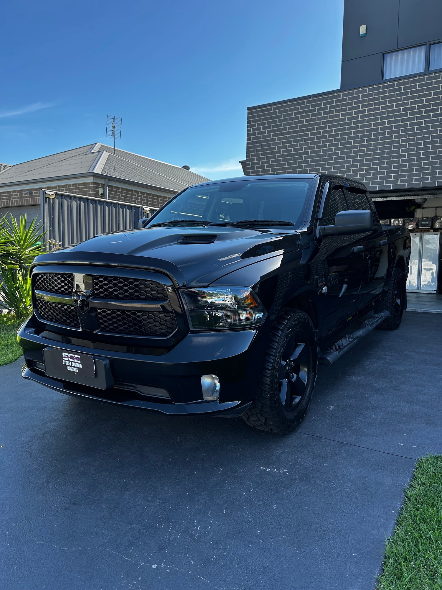 Glossy black RAM pickup truck parked on a residential driveway under clear blue skies, with modern brick and tile houses in the background.
