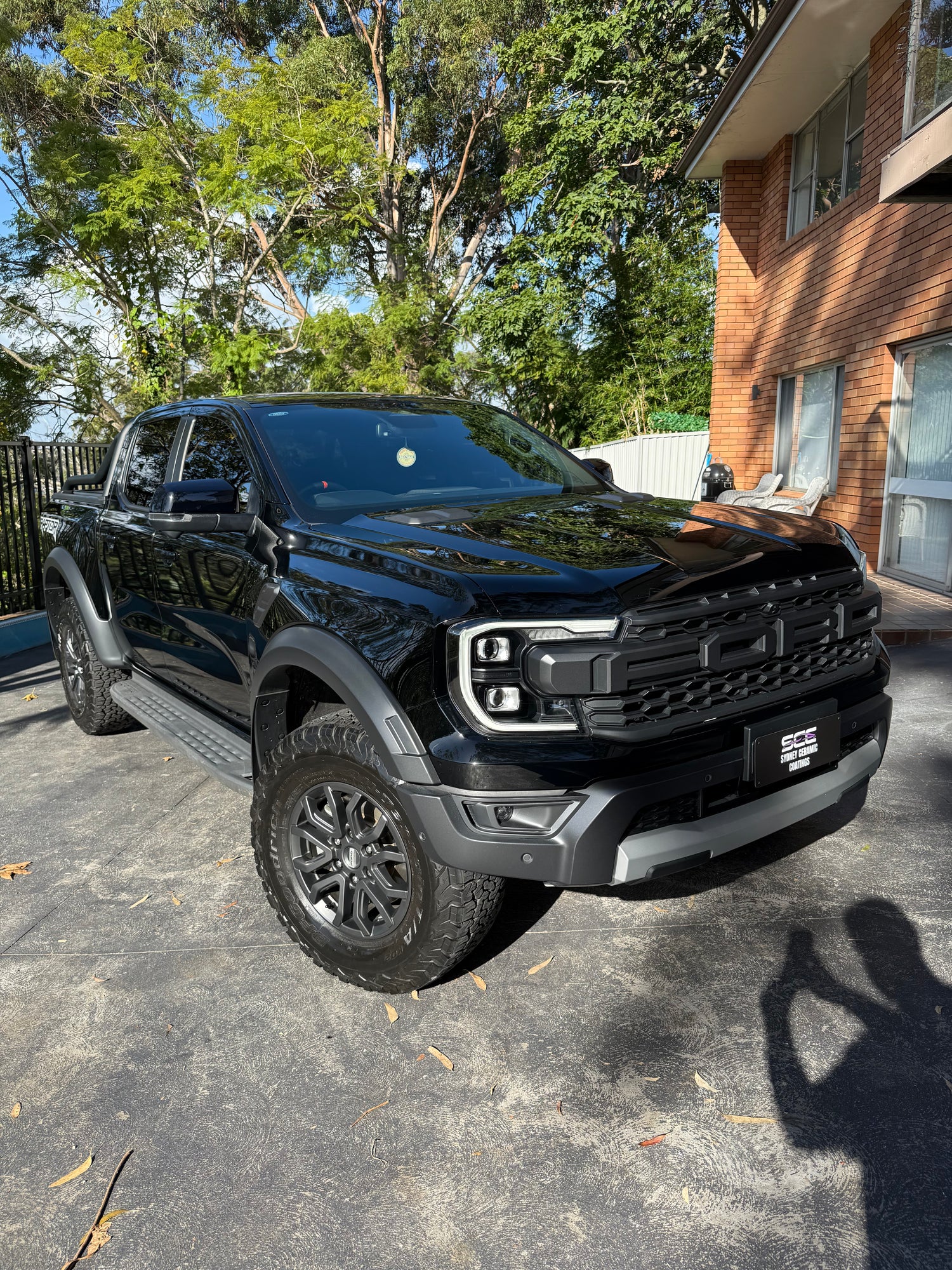 Black Ford Ranger Raptor pickup truck with rugged off-road tires, parked in a sunny residential driveway beside a brick building.