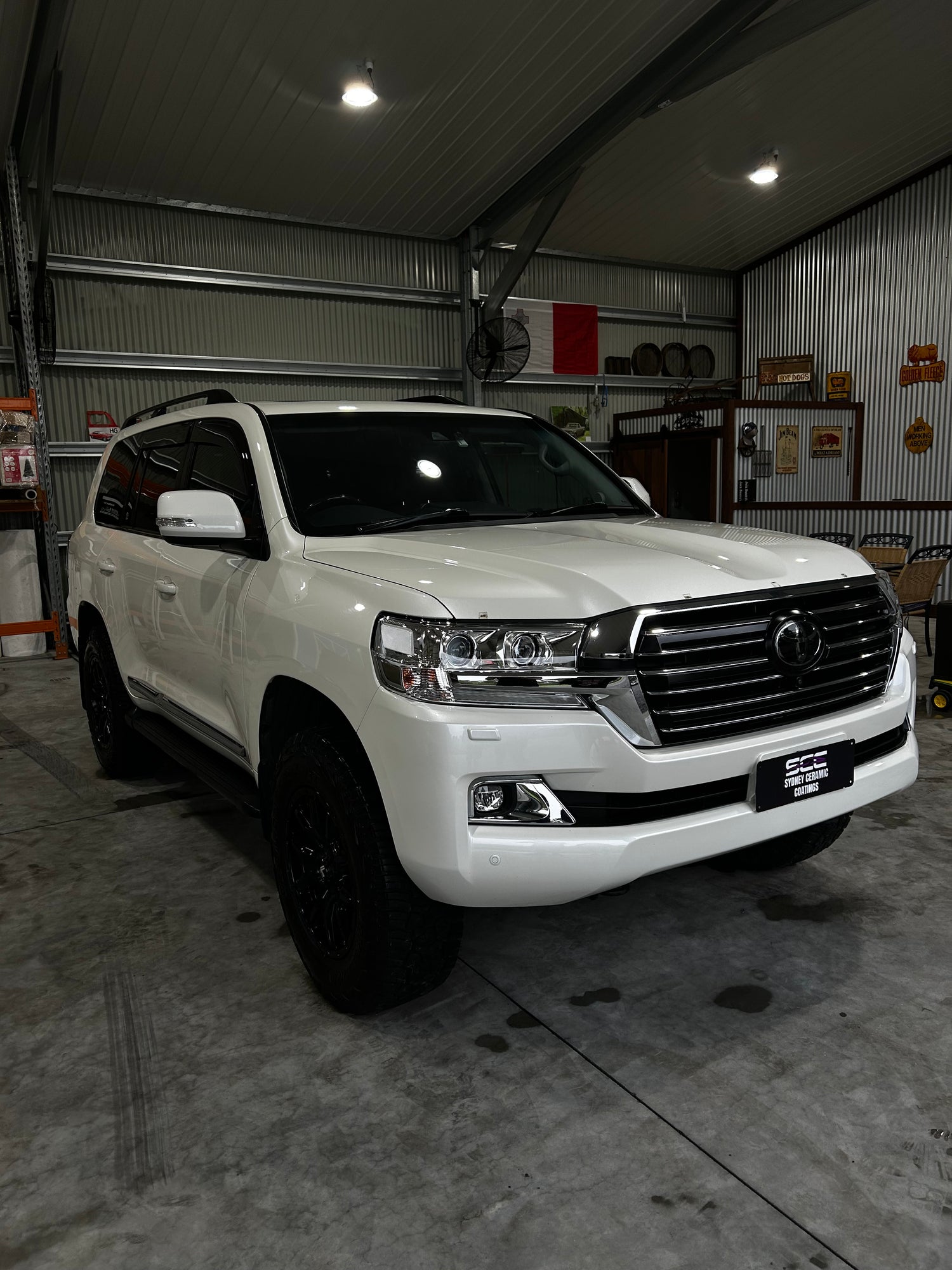 White Toyota Land Cruiser SUV with black wheels parked inside a metal garage, surrounded by workshop tools and vintage signs on the walls.