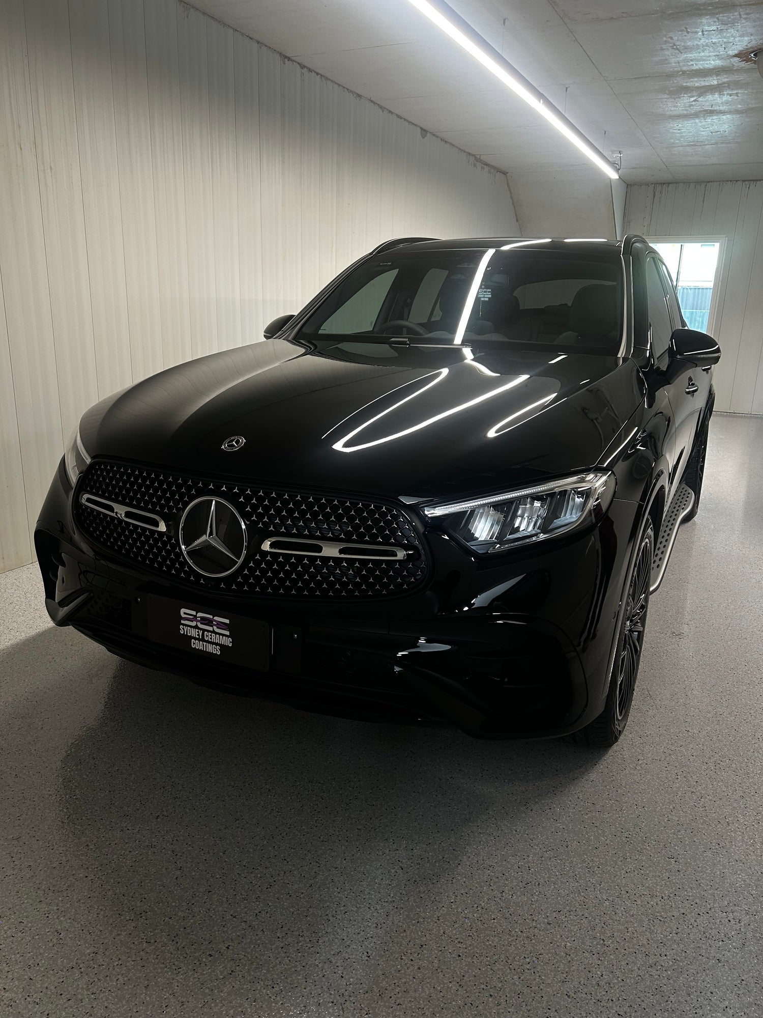Glossy black Mercedes-Benz SUV parked inside a clean, well-lit detailing bay with white walls and epoxy flooring.